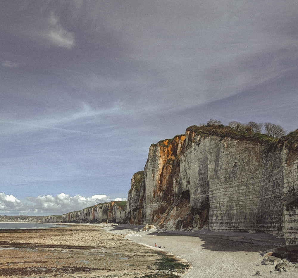 Fotografie van de Imposante krijtrotsen en breed strand in Yport, Normandië, Frankrijk, met een lichte bewolkte lucht en enkele mensen die op het strand wandelen.